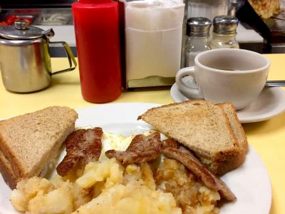 2 Egg platter w Sausage. This picture depicts an order of two eggs Fried with on fried potatoes, sausage and Toast cut in half on top of a platter with a cuppa coffee on the side, served on Johnny’s counter tap