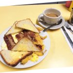2 Eggs Any Style Platter. This picture D pics a platter with scrambled eggs home fried potatoes and sliced toast on top of a white platter served on Johnny’s yellow counter. It also shows a side order of sausage that is usually an additional charge