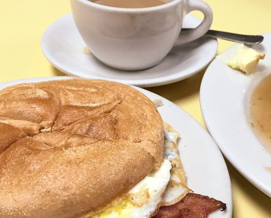 2 Eggs and bacon on a Roll. This picture depicts sandwich that consists of two fried eggs with bacon served on a Kaiser roll that is cut in half on top of a white plate on Johnys yellow countertop