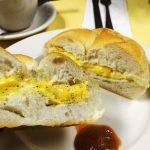 2 Eggs on a Roll w cheese. This picture depicts an egg sandwich that consists of two eggs and American cheese scrambled served on a Kaiser roll that is cut in half on top a white plate Served on Johny’s yellow counter with a cup of coffee on the side