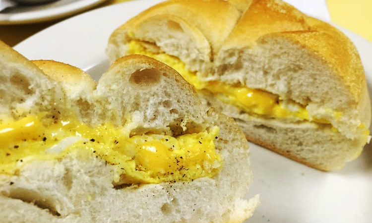 2 Eggs on a Roll w cheese. This picture depicts an egg sandwich that consists of two eggs and American cheese scrambled served on a Kaiser roll that is cut in half on top a white plate Served on Johny’s yellow counter with a cup of coffee on the side