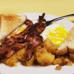 2 egg platter with bacon. This picture depicts two sunny side up eggs Served with home fried potatoes on the side, topped with three pieces of bacon and two slices of toast cut in half placed on top.