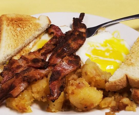 2 egg platter with bacon. This picture depicts two sunny side up eggs Served with home fried potatoes on the side, topped with three pieces of bacon and two slices of toast cut in half placed on top.