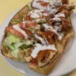 Buffalo Chicken Sandwich. This picture depicts grilled chicken chopped and served on an open faced semolina hero with lettuce tomato spicy hot sauce and ranch dressing drizzled all over served on top of the white plate on Johnny’s