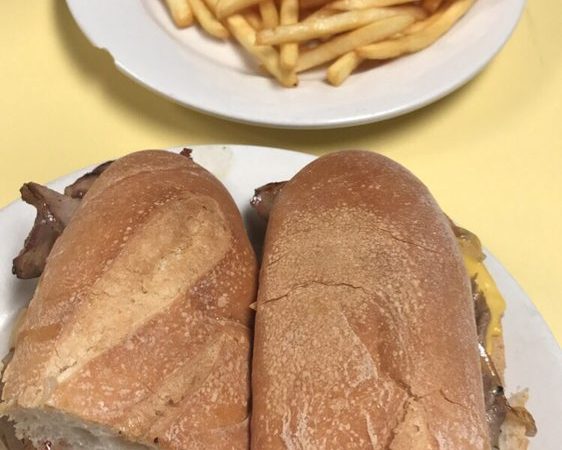 Cheese Steak Hero. This picture depicts a sandwich that consists of sliced steak, peppers and onions, and melted American cheese on a sub that is cut in half served on a white platter accompanied by a side of french fries for an extra charge on top of Johnys counter