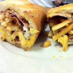 Curious George. This picture shows a sandwich that consists of three Fried eggs, Bacon and sliced ham, Melted American cheese and french fries on a semolina hero that is cut in half and served on a White platter on top of Johnys countertop