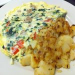 Farmers Omelette. This picture depicts an omelette that is consisted of spinach, mushrooms, tomato and onion with home fried potatoes on the side served on a white plate on Johnny’s counter top