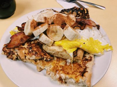 King Kong French toast. This picture depicts French toast made out of eight sub roll that is dipped into the batter. This picture contains the French toast itself topped with Fried eggs, Sliced bananas topped with bacon and ground cinnamon