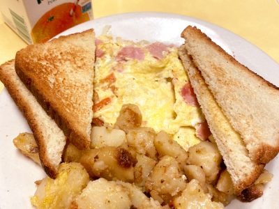 Western Omelette. This picture depicts an omelette which consists of chopped ham, peppers and onions served with home fries potatoes on the side and two slices of toast on top, served on a white planter on Johnny’s counter top