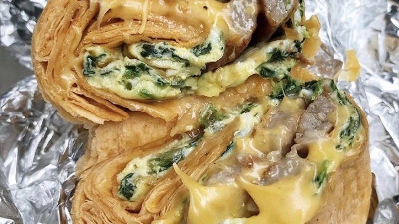 Spinach Egg Sausage and Cheese Wrap. This picture depicts a rap that consists of Two eggs, spinach, sausage and melted cheese that is cut in half ends Served open face on a piece of take out aluminum foil