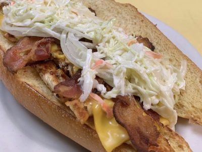 Sloppy Johny. This picture he picks a sandwich on a sub/hero with grilled chicken, raw onion, American cheese, crispy bacon and homemade coleslaw swerved on a white plate on Johnys yellow countertop
